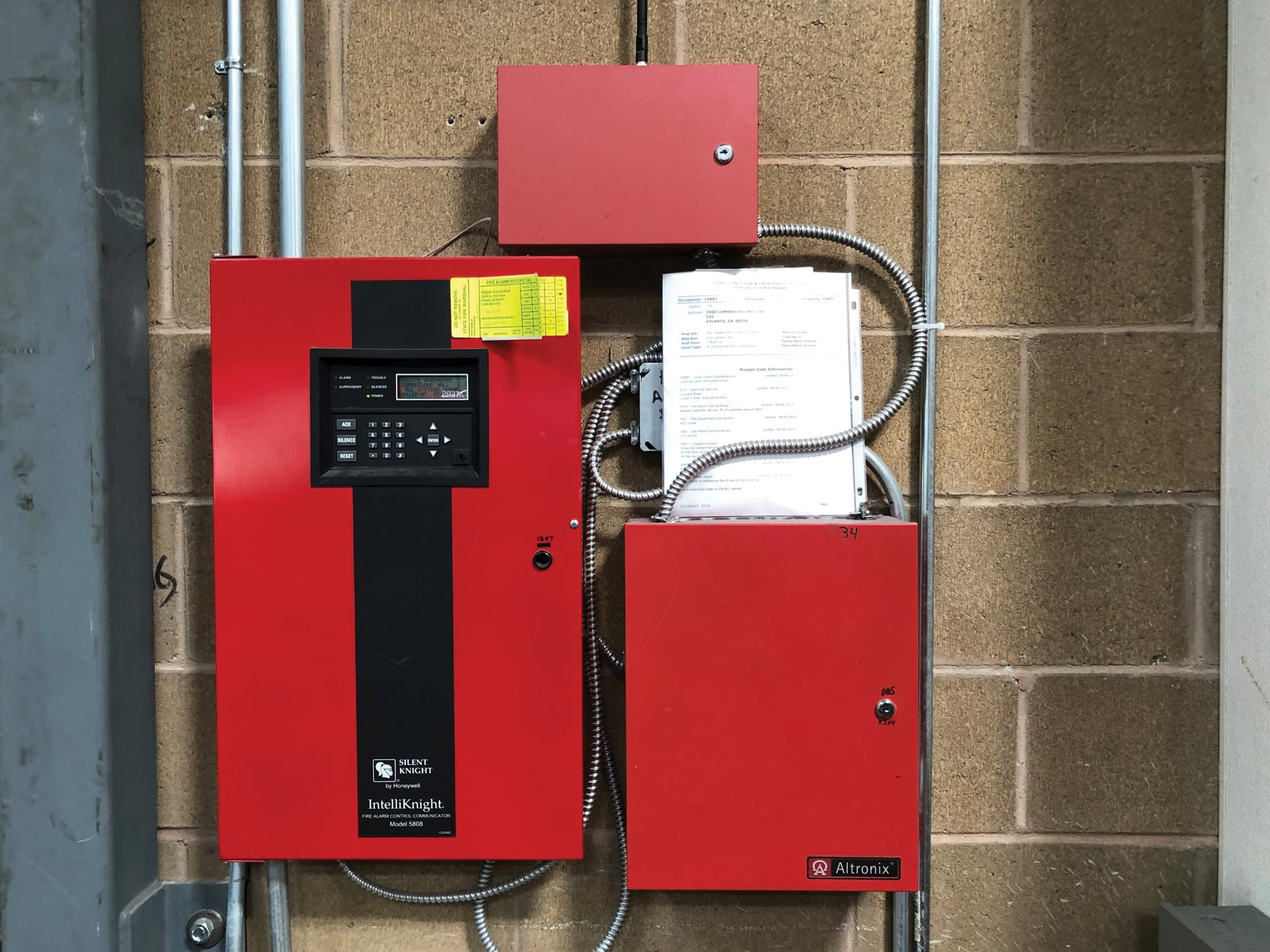 A Telguard cellular communicator is installed above a Silent Knight fire alarm panel and a power supply in a Costco retail store. 
