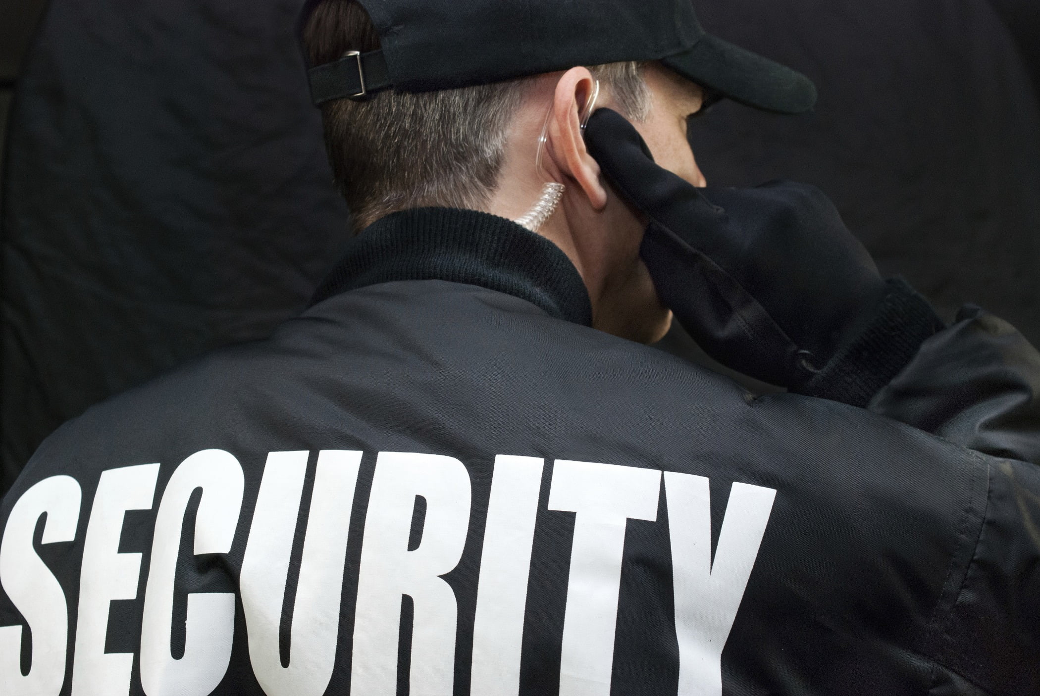 Security Guard Listens To Earpiece, Back of Jacket Showing